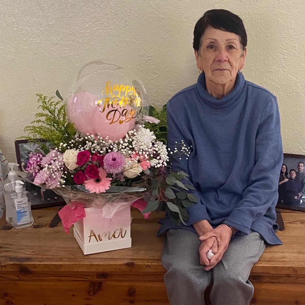Celia Márquez sits on a wooden bench. A bouquet of pink and white flowers with a balloon is beside her. She is wearing a cornflower blue sweater and grey pants.