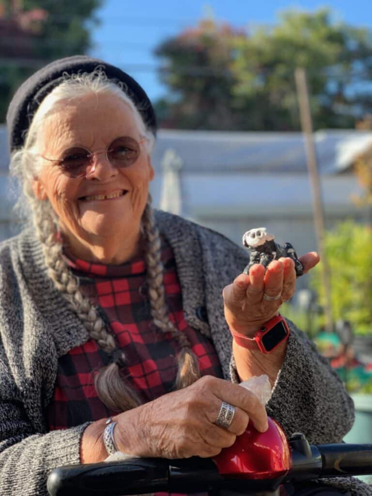 A photo of Victoria. She is smiling and wearing a grey sweater and red checkered shirt. She is wearing silver rings, and holding a figurine in her left hand.