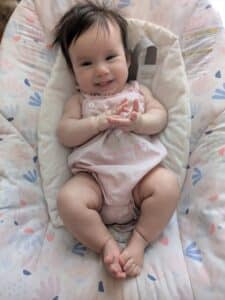 A photo of Baby Valentina taken from above, she is smiling and resting on a soft blanket. She is wearing a pink onesie that reads "Daddy's Little Girl."