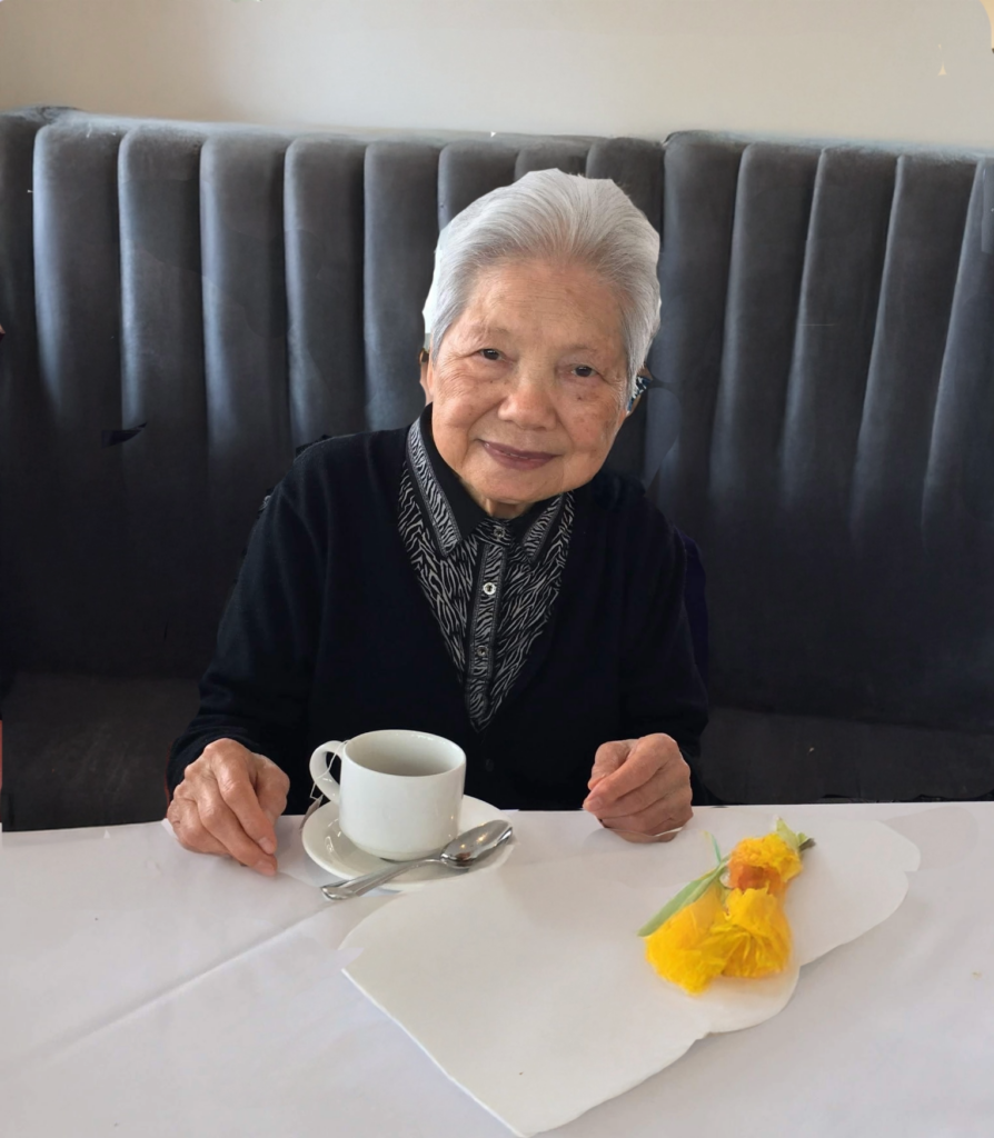 Gugu is seated at a restaurant booth with a white mug of tea and yellow flowers resting in front of her on the table. She is wearing a black cardigan sweater over a zebra-print button-up shirt.